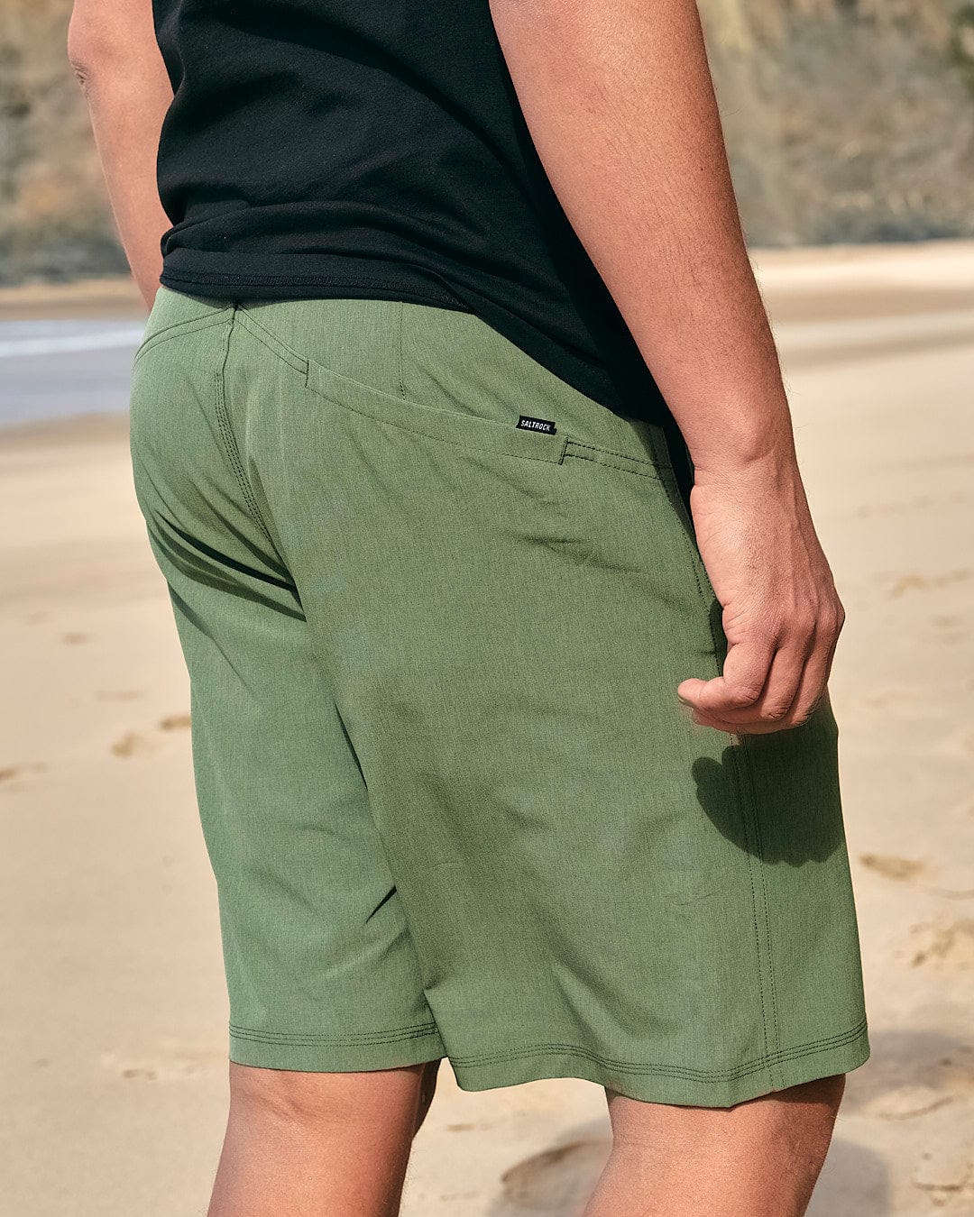 An essential man's wear, the Saltrock Cargo Amphibian II - Mens Boardshort - Dark Green are donned by a man standing on the beach.