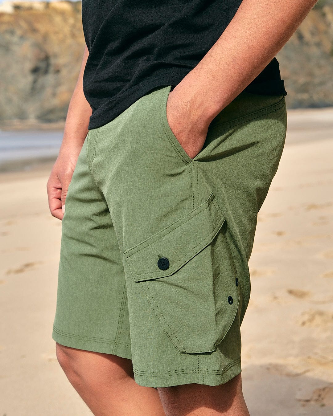 A man is standing on the beach wearing green Saltrock Cargo Amphibian II - Mens Boardshort - Dark Green shorts.