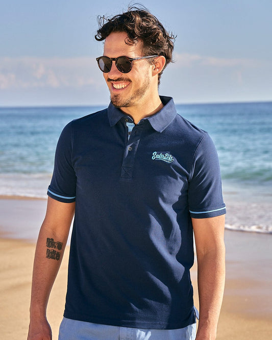A man sporting a cool Saltrock Roland - Mens Short Sleeve Polo Shirt - Blue on the beach.
