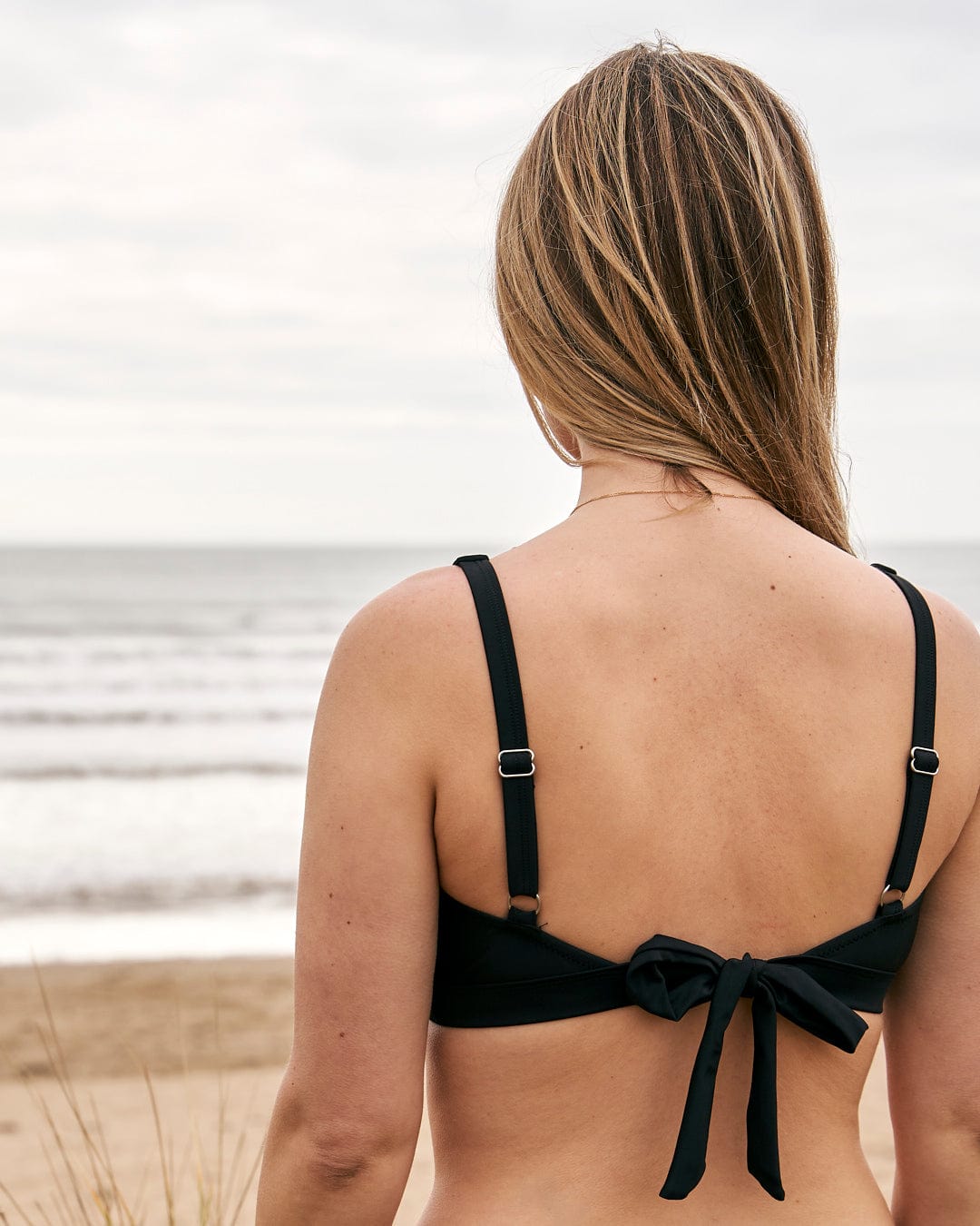 The woman is pictured from the back, wearing a Saltrock Rosie - Womens Bikini Top - Black featuring adjustable straps.