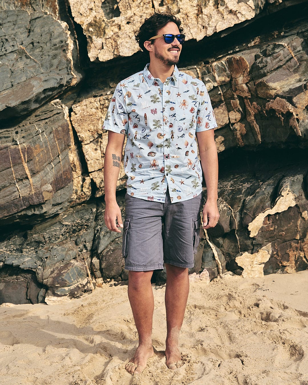A man standing on the beach wearing Saltrock sunglasses and a Ruan - Mens Washed Short Sleeve Shirt - Light Blue.