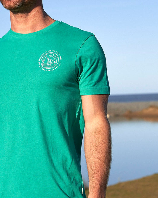 A man wearing a Saltrock Sailaway Outline - Mens Short Sleeve T-Shirt - Bright Green standing in front of a lake.