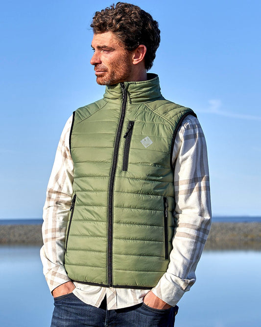 A man in a green vest, wearing the Saltrock - Mens Reversible Gilet, standing by the water during an outdoor activity.