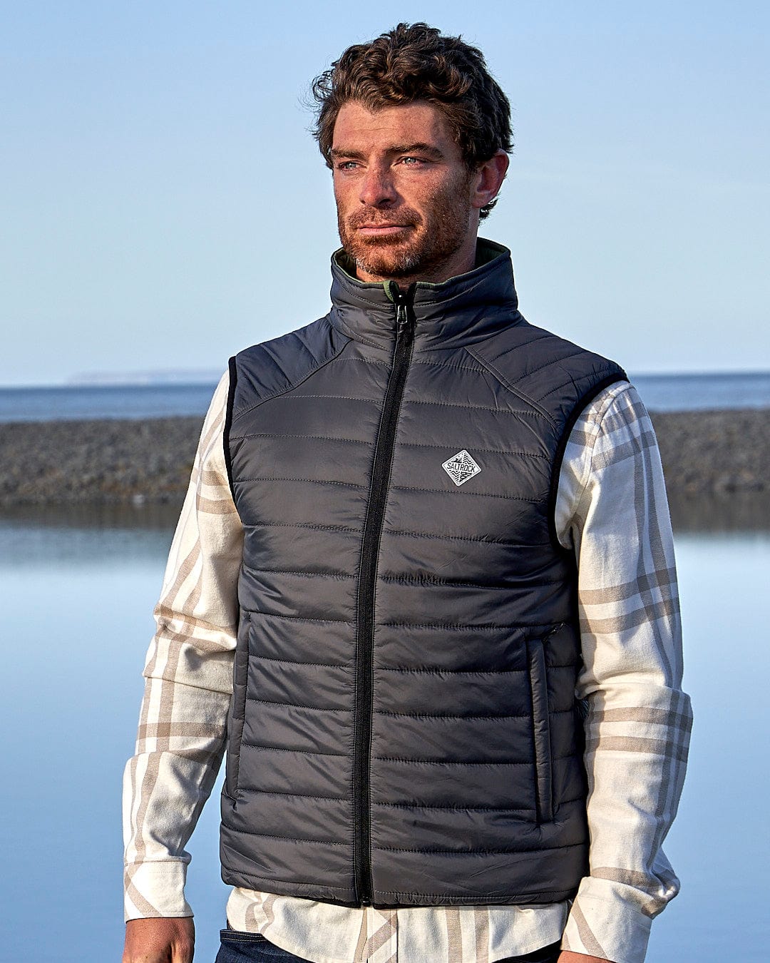 A man standing in front of a body of water wearing a Saltrock - Mens Reversible Gilet - Dark Green/Black.