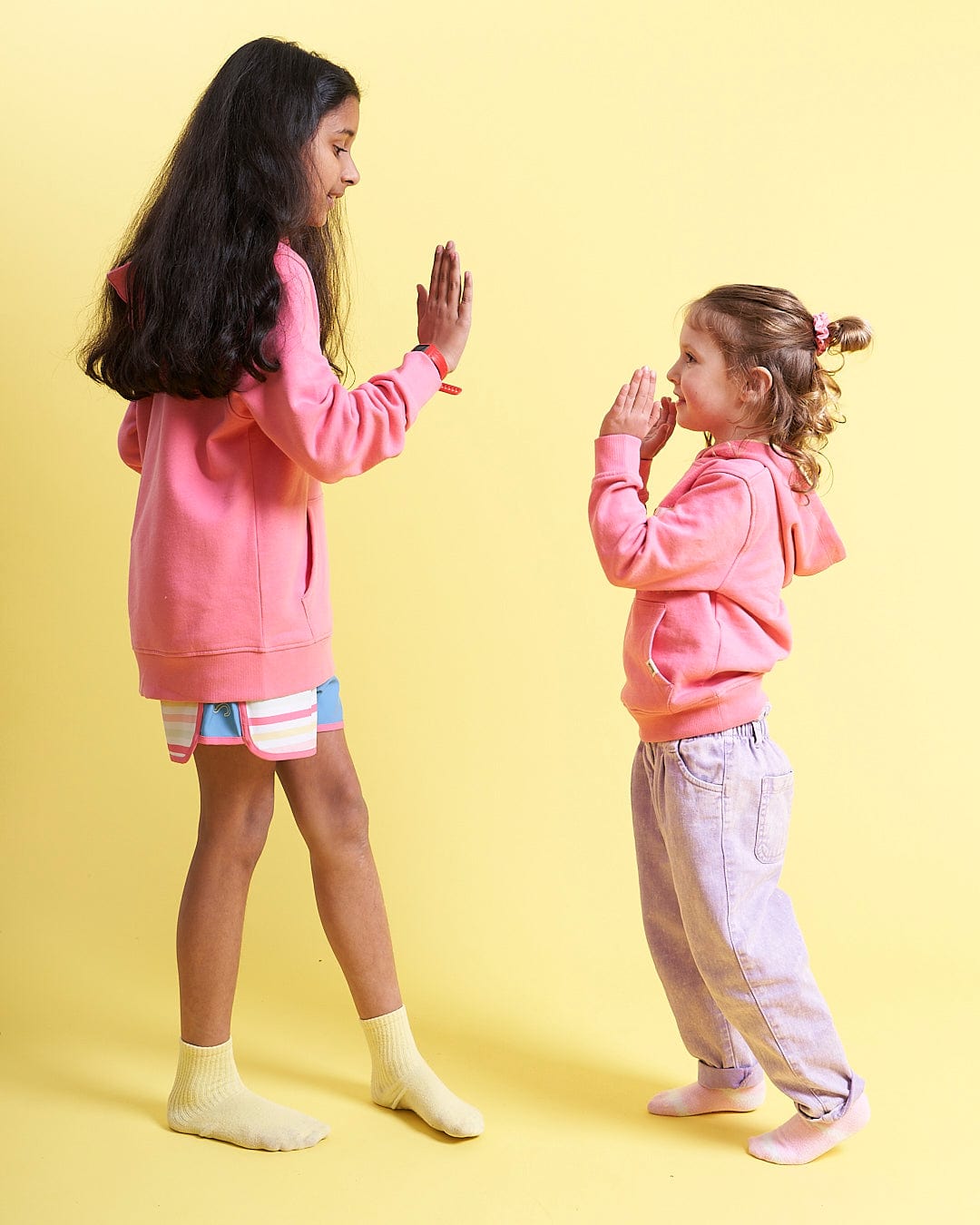 Two little girls in Seabed - Kids Pop Hoodie - Pink sweatshirts standing on a yellow background.