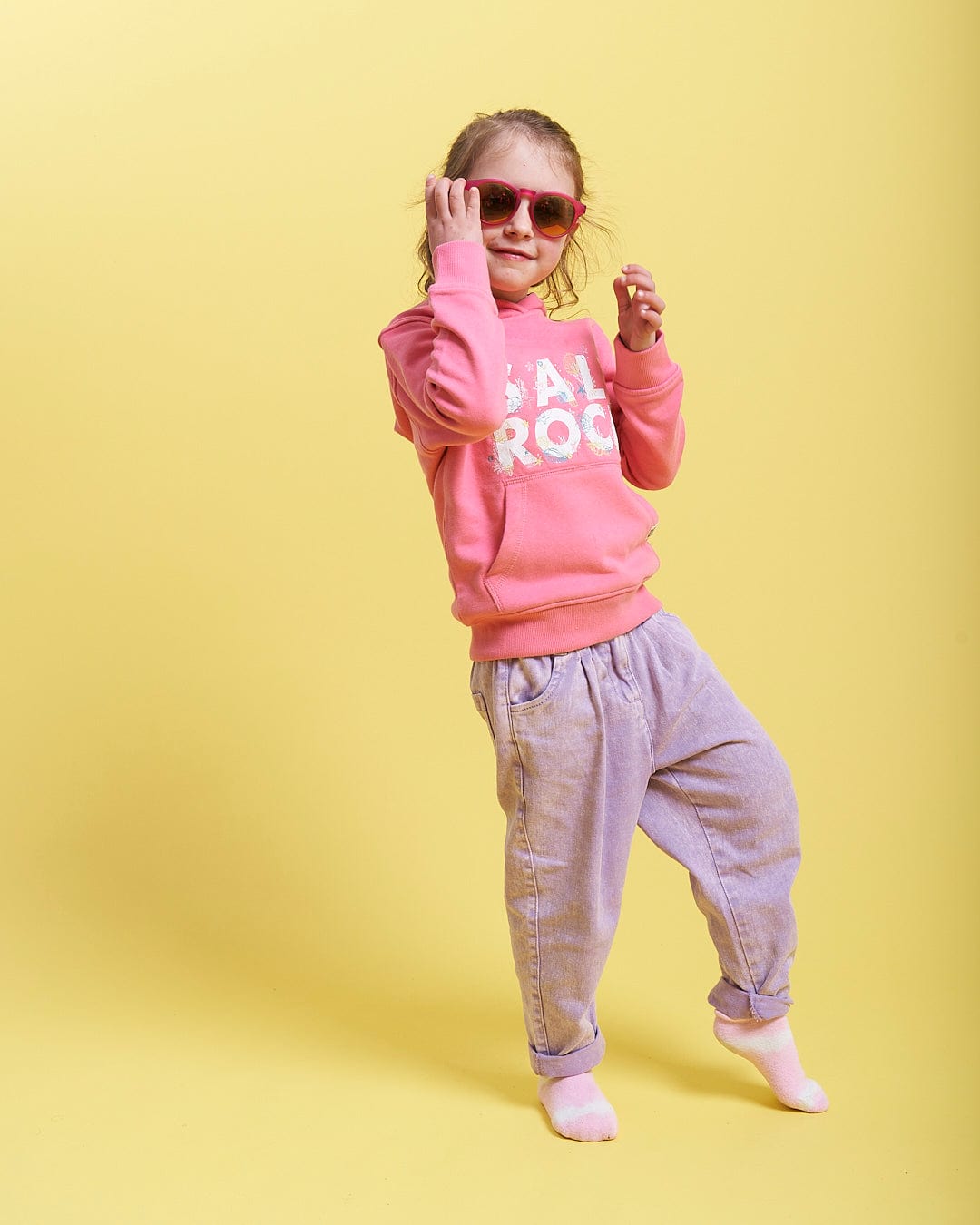 A girl wearing Saltrock sunglasses and a Seabed - Kids Pop Hoodie - Pink sweatshirt on a yellow background.