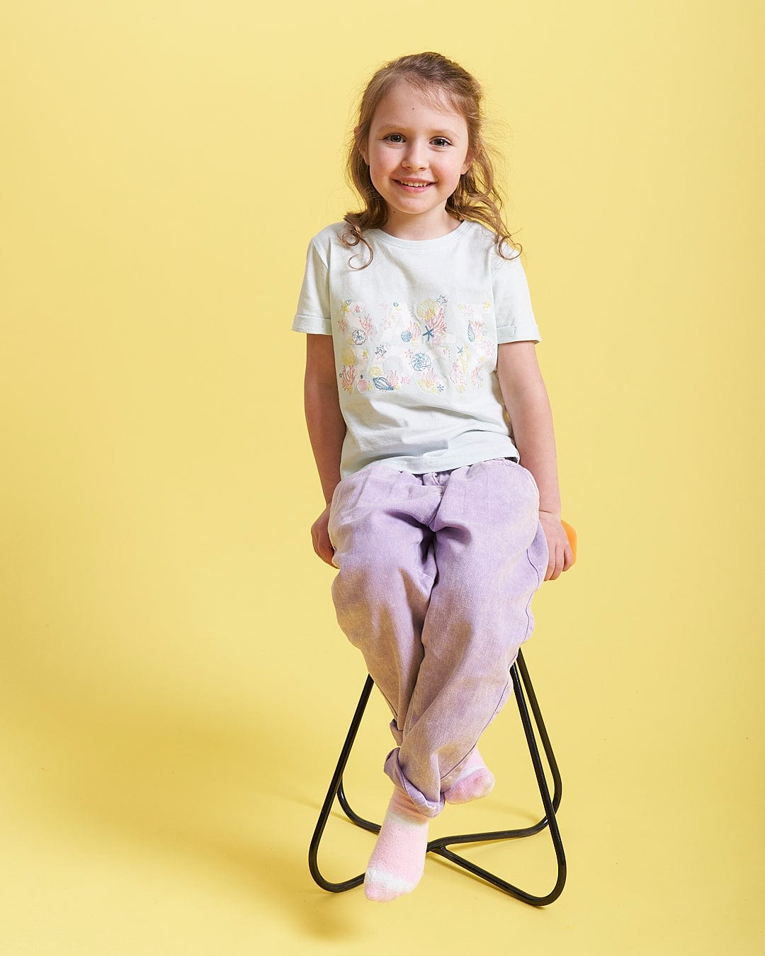 A little girl sitting on a stool wearing a Saltrock Seabed - Kids Short Sleeve T-Shirt - Light Blue.