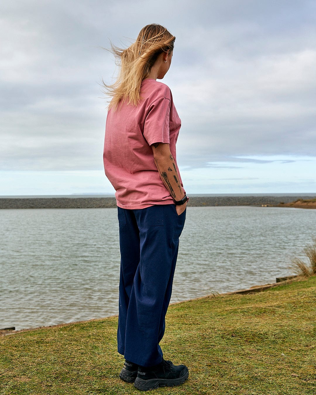 A woman is standing in front of a body of water wearing the Saltrock Sea Shells - Womens Relaxed Fit T-Shirt - Mid Pink.