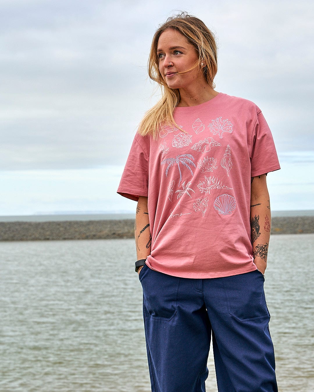 A woman wearing a Saltrock Sea Shells - Womens Relaxed Fit T-Shirt - Mid Pink standing by the water.