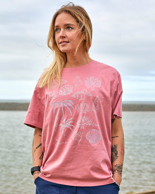 A woman wearing a Saltrock Sea Shells - Womens Relaxed Fit T-Shirt - Mid Pink standing by the water.