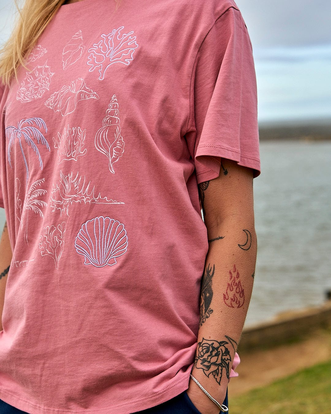 A woman wearing a Saltrock Sea Shells - Womens Relaxed Fit T-Shirt - Mid Pink near the water.