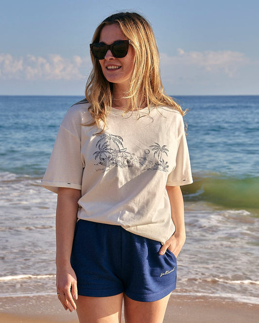 A woman wearing a Sea View - Womens Boyfriend Fit T-Shirt - White by Saltrock and blue shorts standing on the coastal beach.
