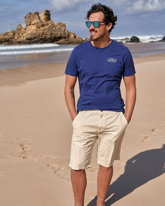 A man standing on a beach wearing a blue t-shirt and Saltrock's Sennen - Mens Chino Short - Natural.