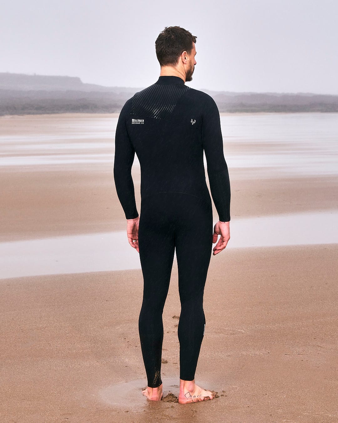 A man in a Saltrock Shockwave - Mens 3/2 Chest Zip Full Wetsuit - Black standing on the beach.