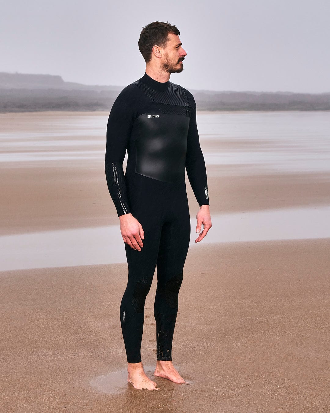 A man standing on the beach in a Saltrock Shockwave - Mens 3/2 Chest Zip Full Wetsuit - Black.