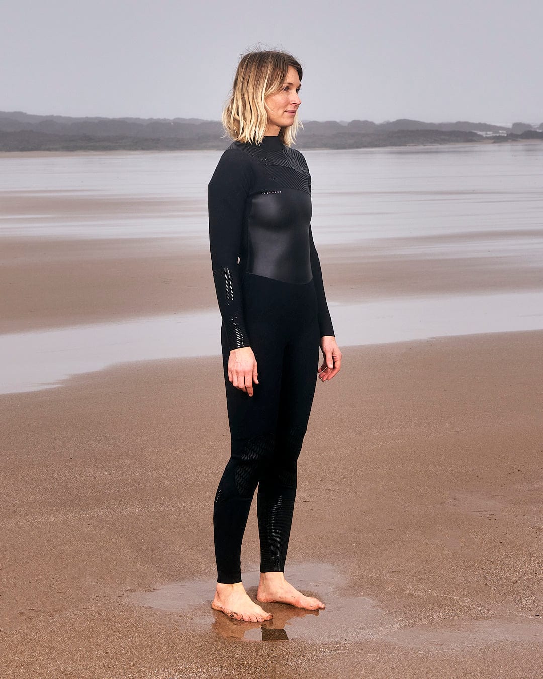 A woman in a Saltrock Shockwave - Womens 3/2 Front Zip Full Wetsuit - Black standing on the beach.