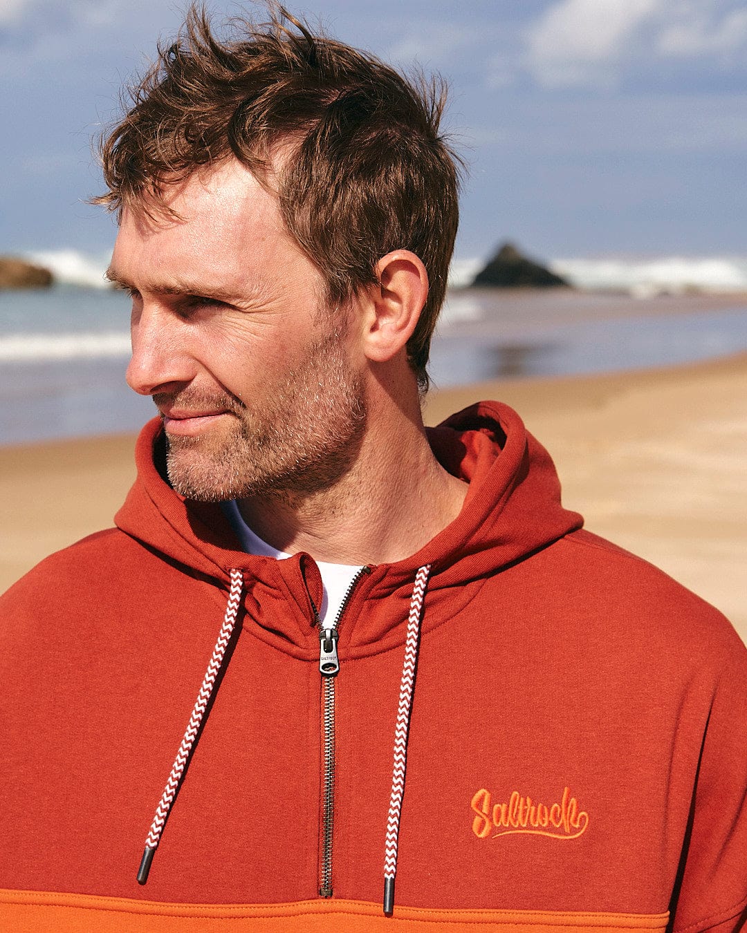 A man is standing on the beach wearing a Saltrock Speed Embroidery - Mens 1/4 Neck Zip - Red hoodie.