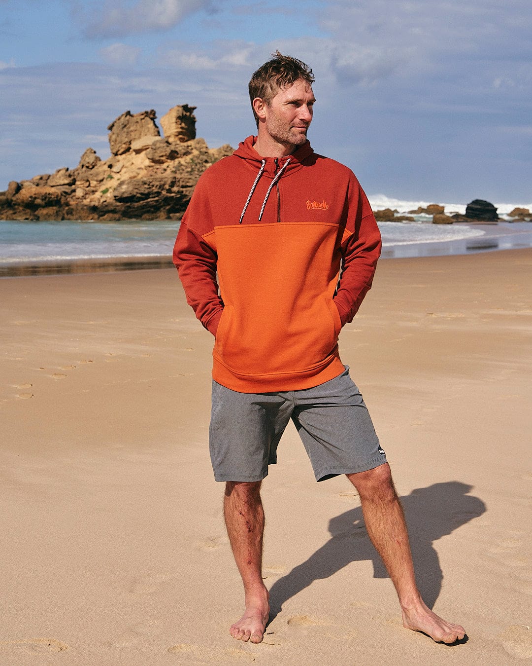 A man in a Saltrock Speed Embroidery - Mens 1/4 Neck Zip - Red sweatshirt standing on a beach.
