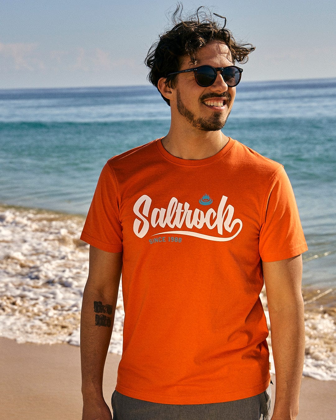 A man wearing sunglasses and a Speed - Mens Short Sleeve T-Shirt - Red by Saltrock standing on the beach.