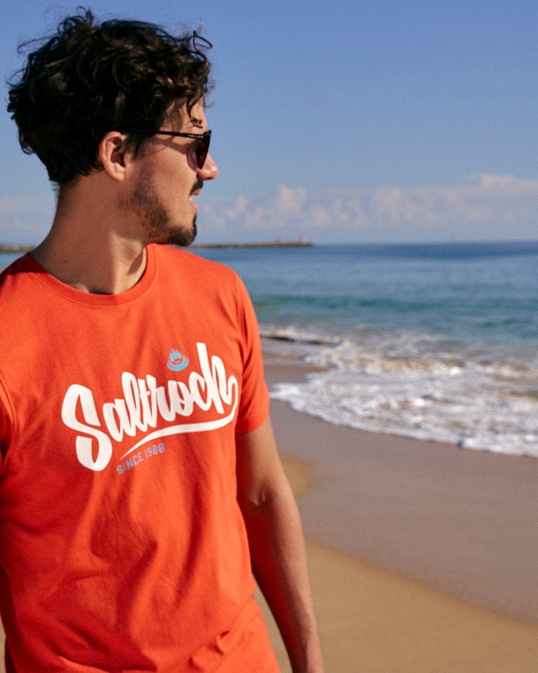 A man wearing sunglasses and a Saltrock Speed-Mens Short Sleeve T-Shirt - Red standing on the beach.