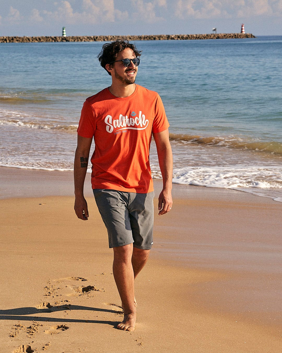 A man walking on a beach near the ocean wearing a Saltrock Speed - Mens Short Sleeve T-Shirt - Red.