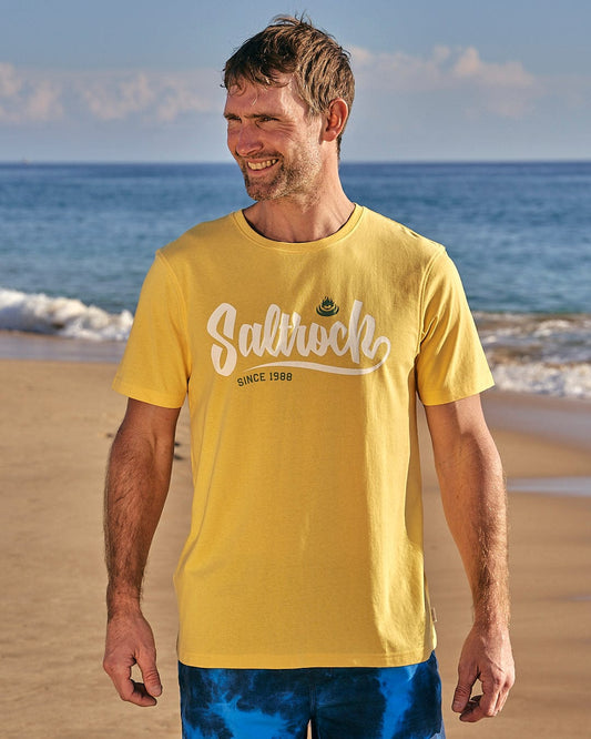 A man is standing on the beach wearing a Saltrock Speed - Mens Short Sleeve T-Shirt - Yellow.