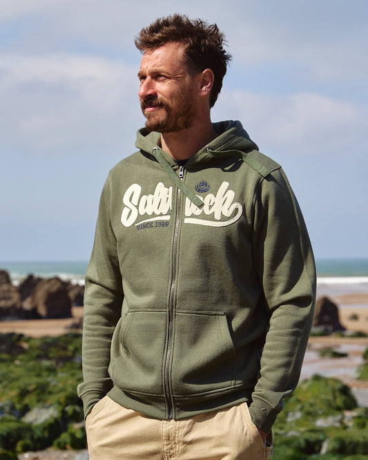 A man standing on a beach wearing a Saltrock Speed - Mens Zip Hoodie - Dark Green.