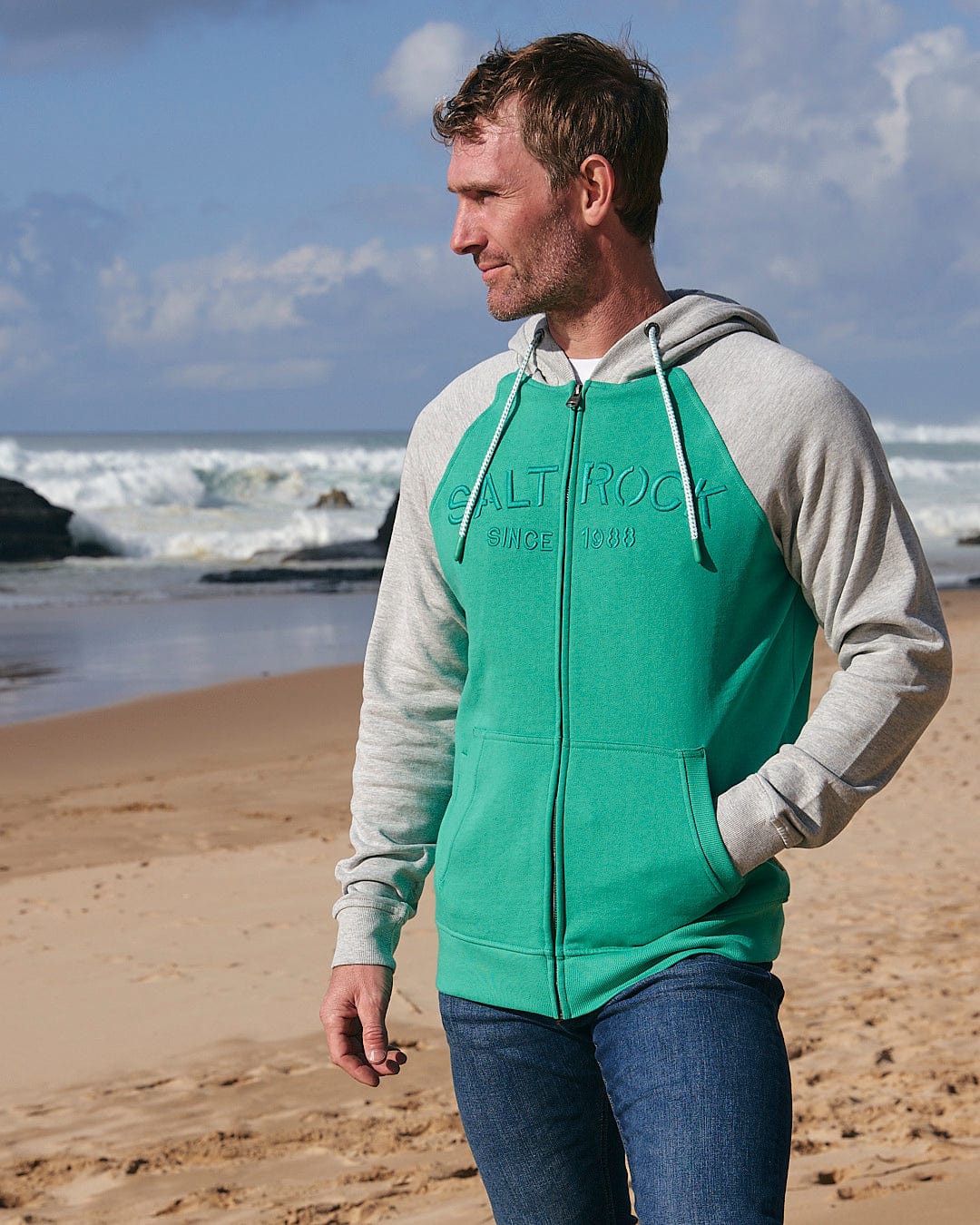 A man standing on a beach wearing a Saltrock Stencil - Mens Zip Hoodie - Green.