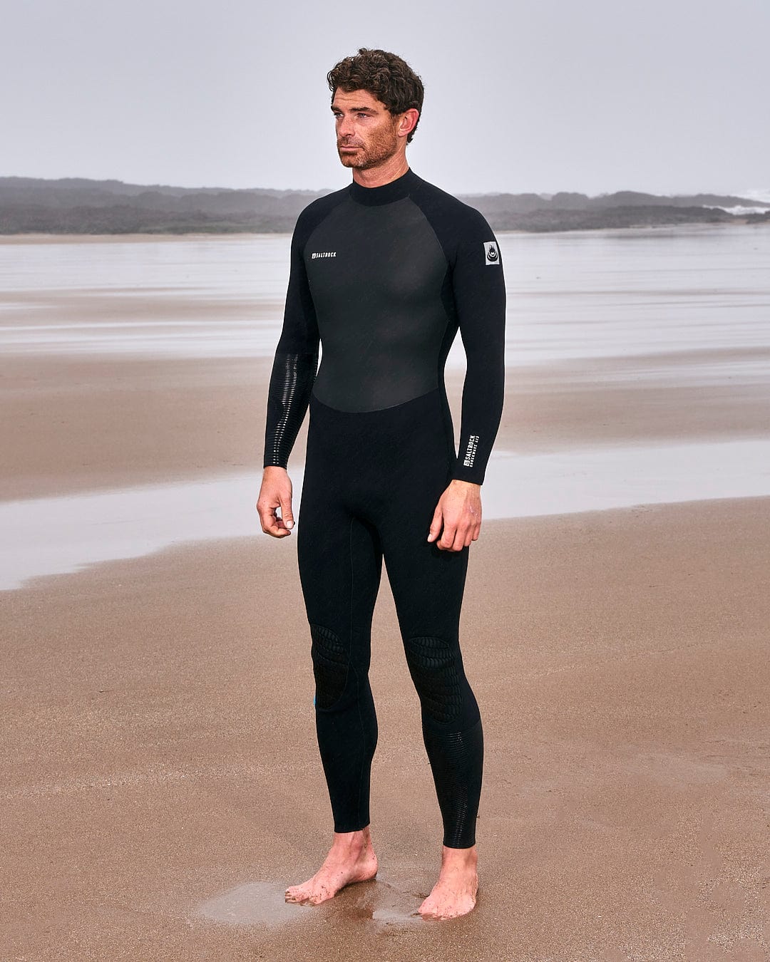 A man standing on the beach in a Saltrock Synthesis - Mens 4/3 Back Zip Full Wetsuit - Black.