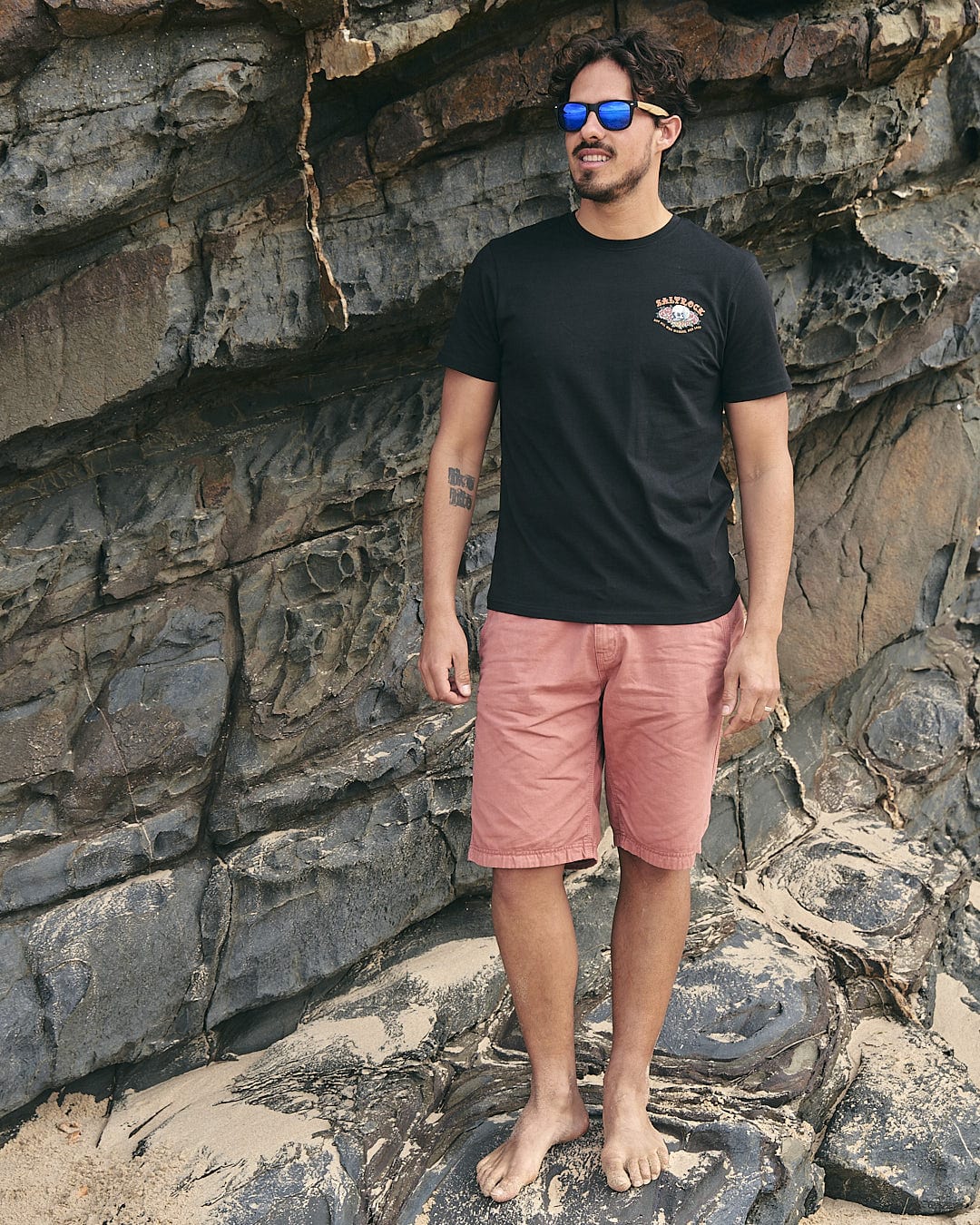 A man wearing sunglasses and a Saltrock - Tattoo Island - Mens Short Sleeve T-Shirt - Black standing on rocks.