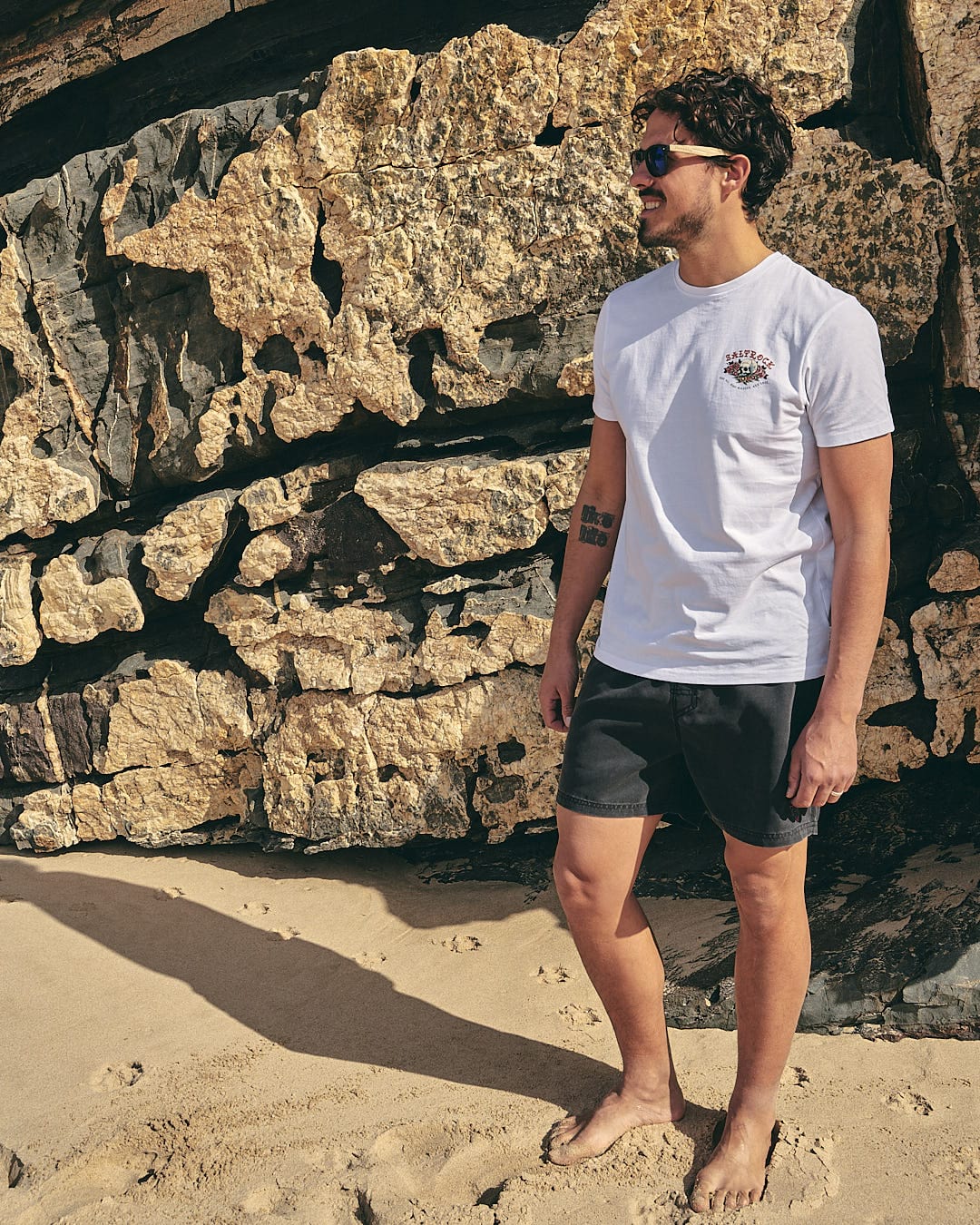 A man standing on the beach wearing a Saltrock - Tattoo Island Mens Short Sleeve T-Shirt in White and shorts.