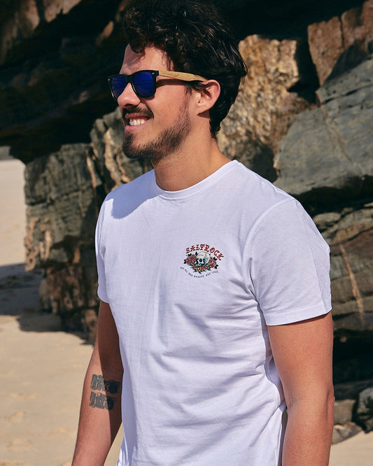 A man wearing sunglasses and a Saltrock - Tattoo Island Mens Short Sleeve T-Shirt - White on a beach.