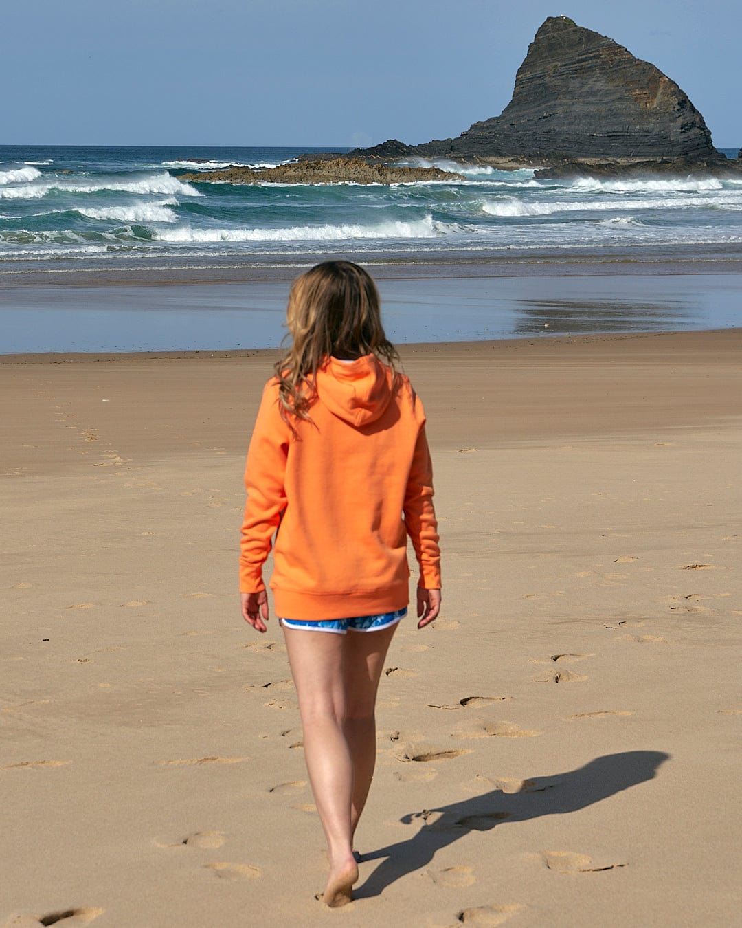 A girl in a Saltrock Velator - Womens Pop Hoodie - Light Orange walking on the beach.