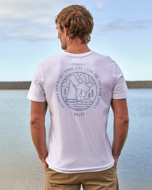 A man standing by the water wearing a Saltrock Wales Sailaway Outline - Mens Short Sleeve T-Shirt - White.
