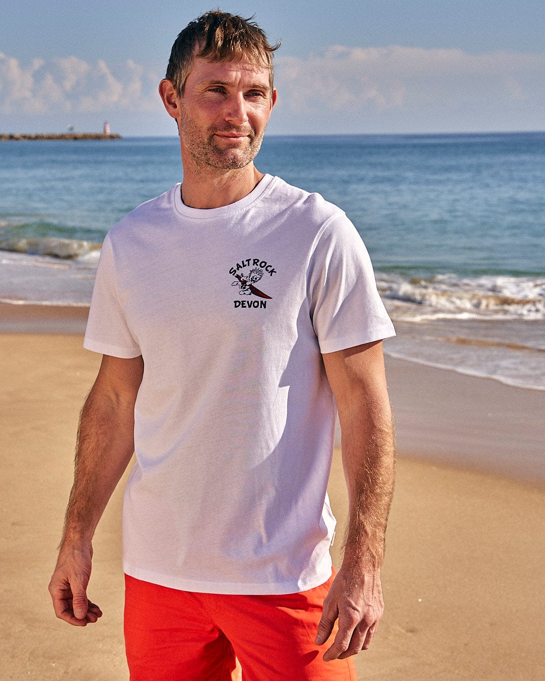 A man is standing on the beach wearing a Saltrock Wave Rider Devon - Mens Short Sleeve T-Shirt - White.