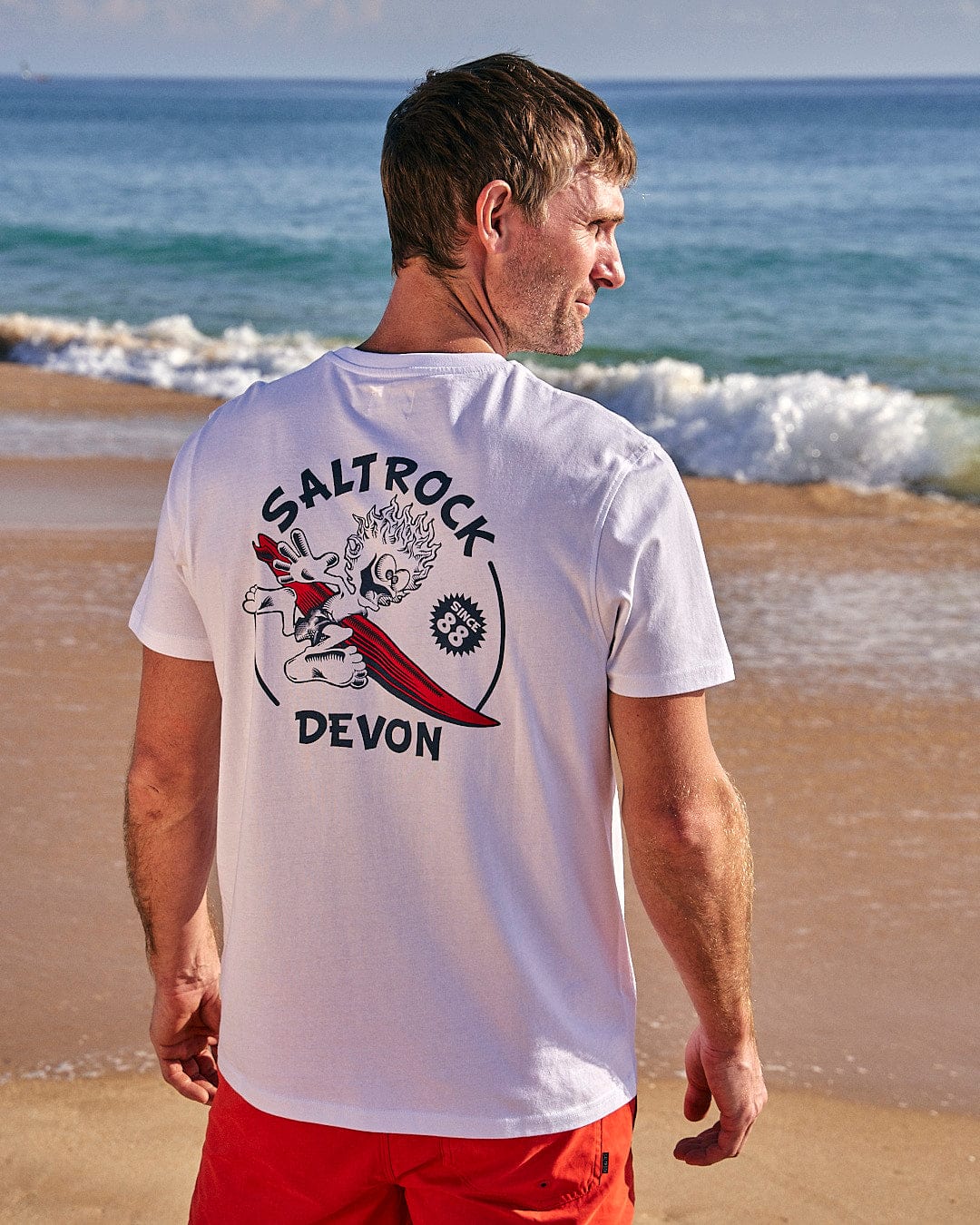 A man is standing on the beach wearing a Saltrock Wave Rider Devon - Mens Short Sleeve T-Shirt - White.
