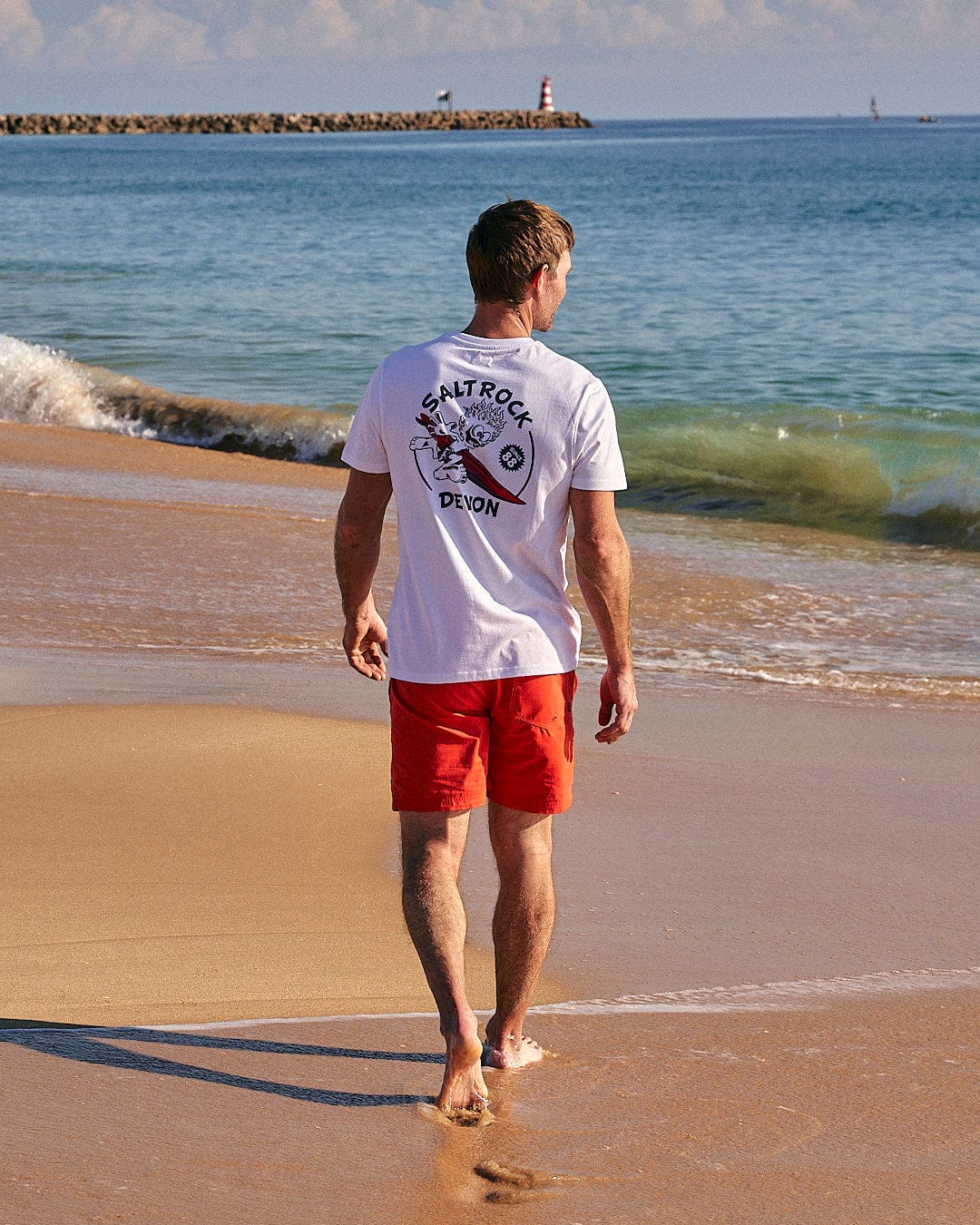 A man walking on a beach with a Saltrock Wave Rider Devon - Mens Short Sleeve T-Shirt - White surfboard.