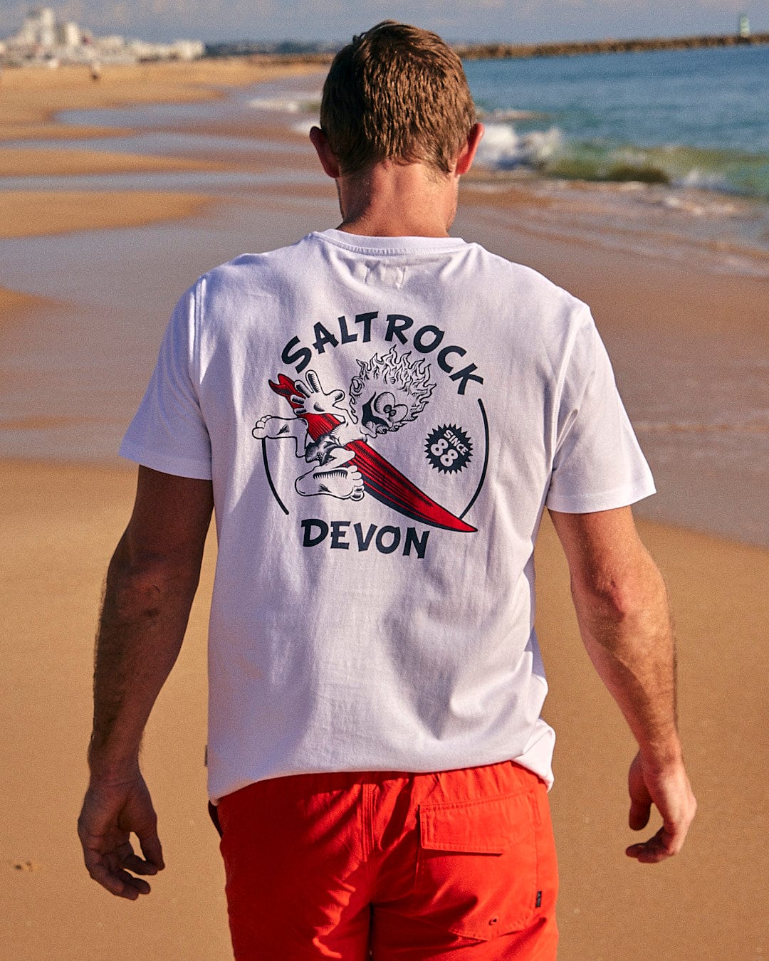 A man walking on the beach wearing a white t - shirt that says Saltrock Wave Rider Devon - Mens Short Sleeve T-Shirt - White.