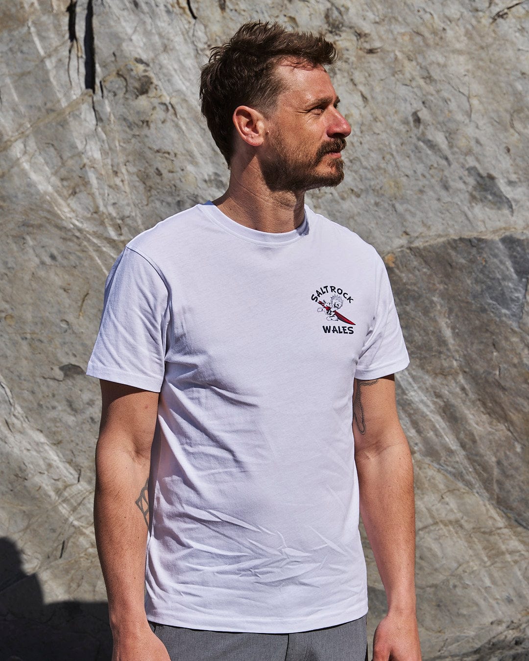 A man wearing a Wave Rider Wales - Mens Short Sleeve T-Shirt in white standing in front of a rock with Saltrock branding.