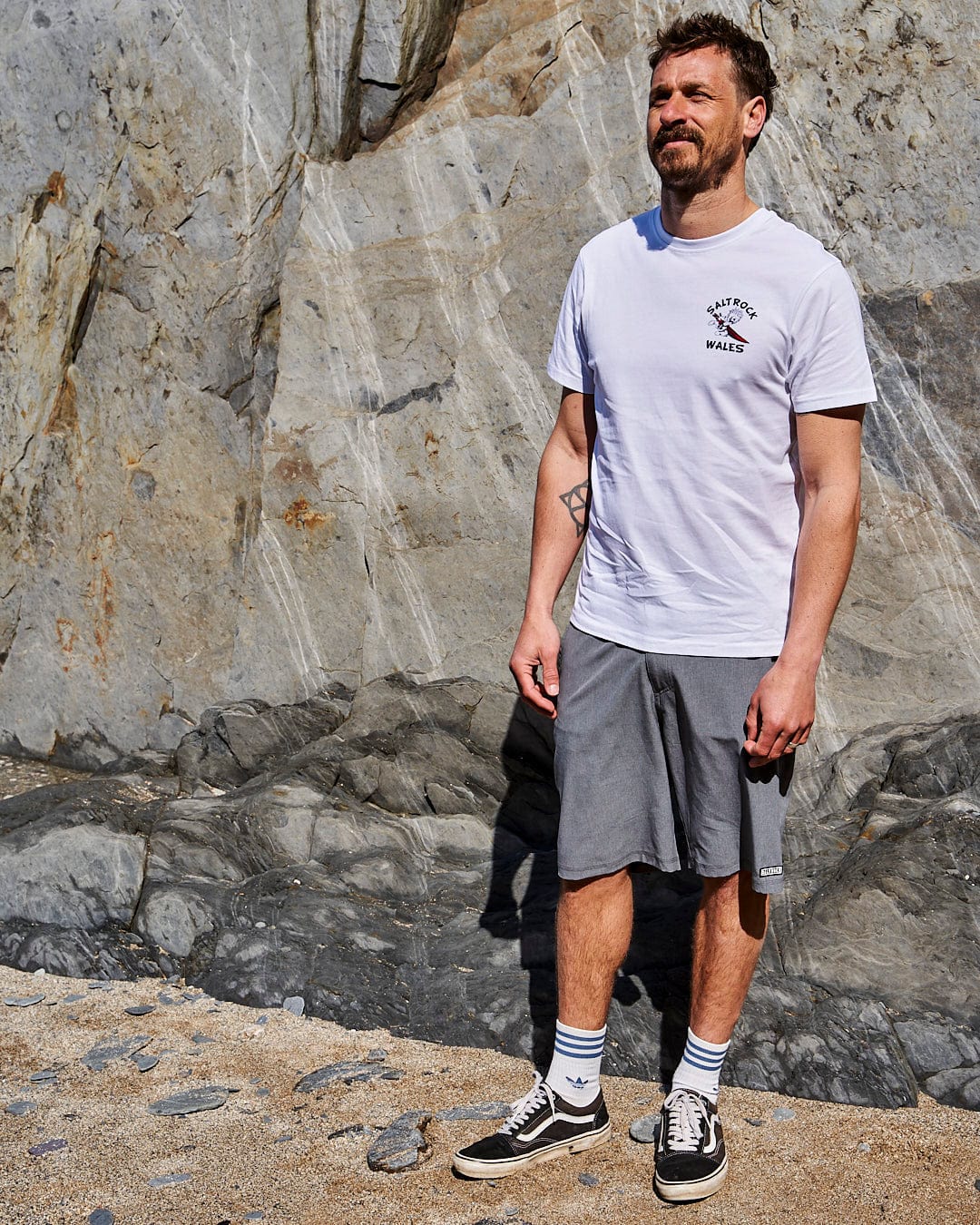 A man wearing a Wave Rider Wales - Mens Short Sleeve T-Shirt in White, standing in front of a Saltrock.